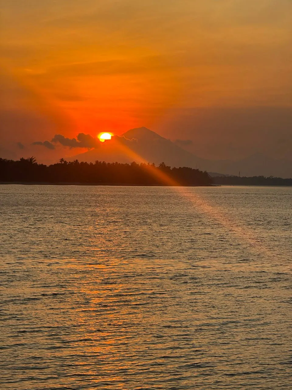The sun stands as an orange disc above a dark strip of coast, behind it the cone of a volcano, a ray of light falls across the water