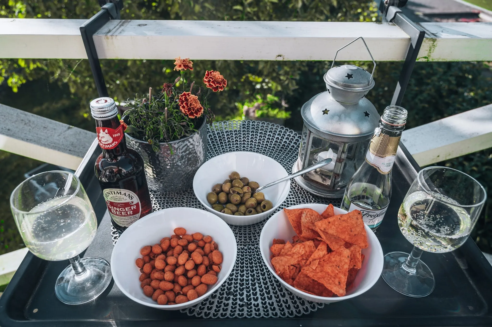 Table with ginger beer, two glasses and bowls of nuts, olives and chips, behind them a balcony railing and a flower pot