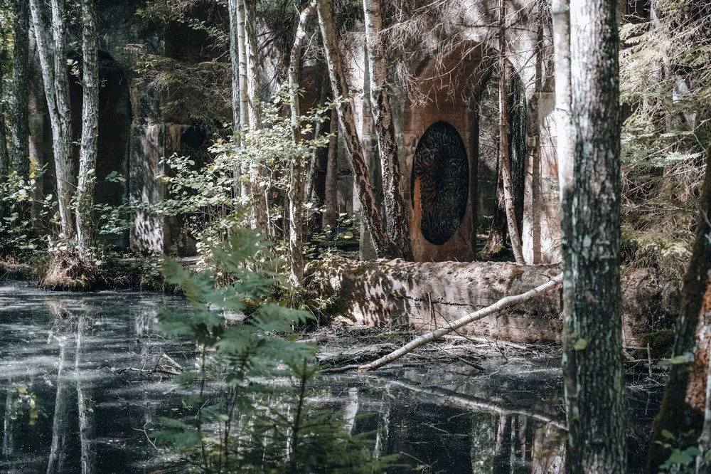 Betonbogen mit runder dunkler Öffnung im Wald, davor Wasser und Birkenstämme