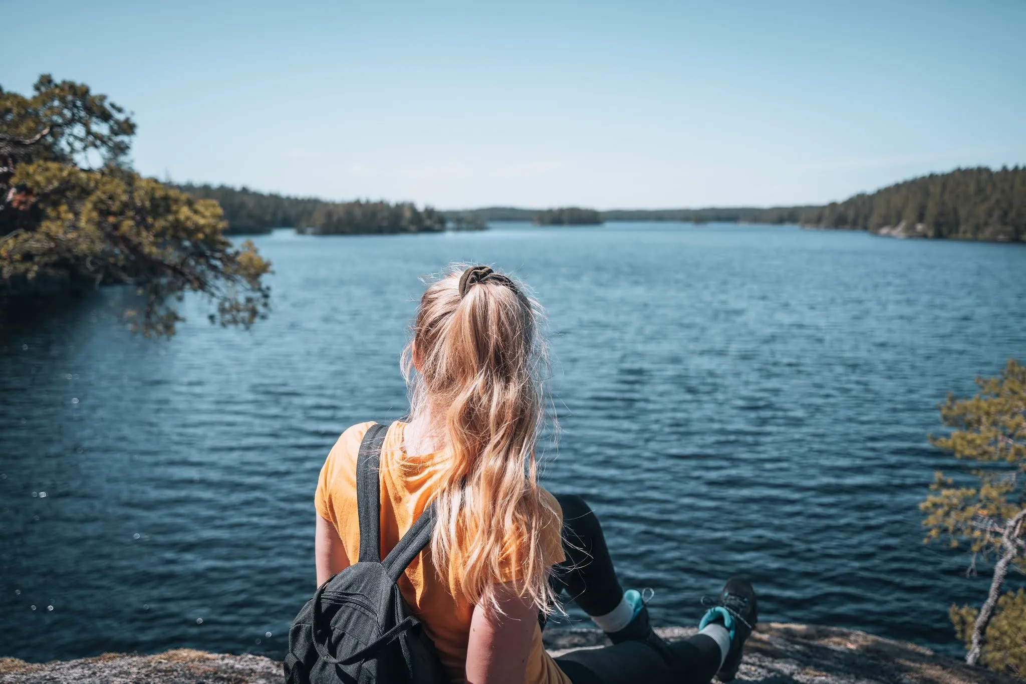 Woman with a backpack sits on a rock in Tiveden National Park and looks out over a lake with wooded shores