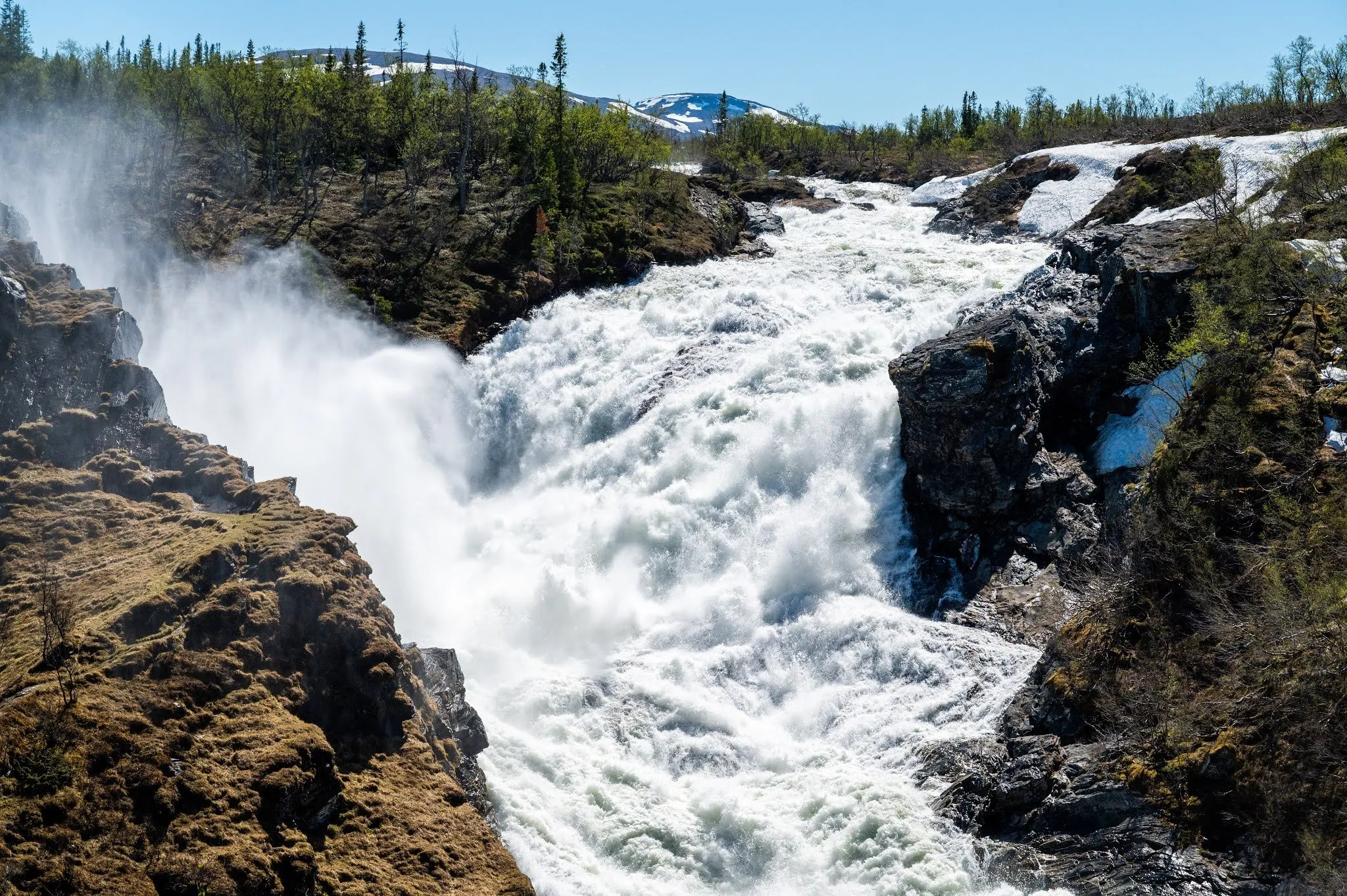White foaming water plunges through a rock gorge, mountains with remnants of snow under a blue sky in the background
