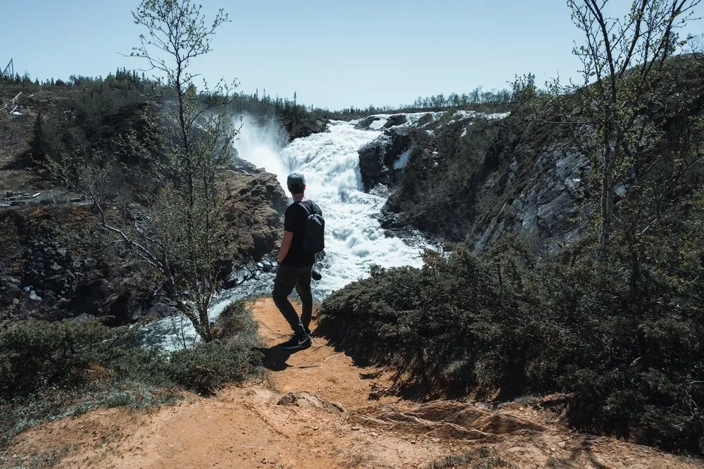 Ein Mann mit Rucksack steht auf dem Pfad zum Aussichtspunkt und blickt auf die Wassermassen des Handölsforsen