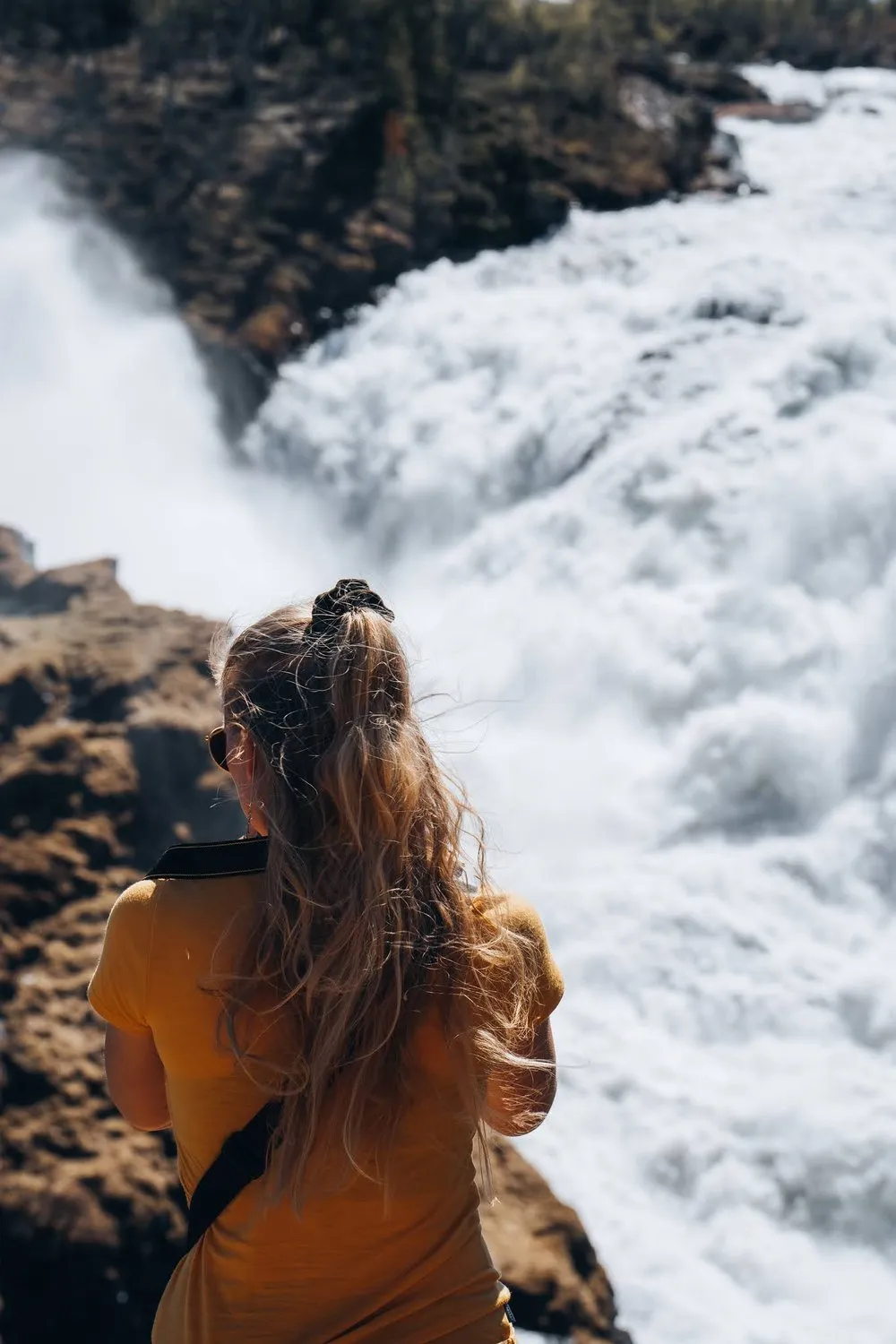 Eine Frau mit hochgesteckten Haaren schaut, von hinten aufgenommen, von einem Felsen auf das weiss schäumende Wasser des Handölsforsen