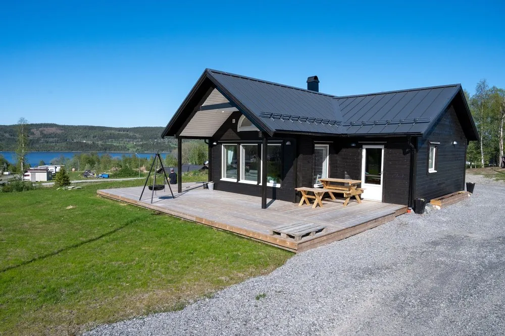 Das moderne schwarze Holzhaus in Huså mit grosser Holzterrasse, im Hintergrund ein See