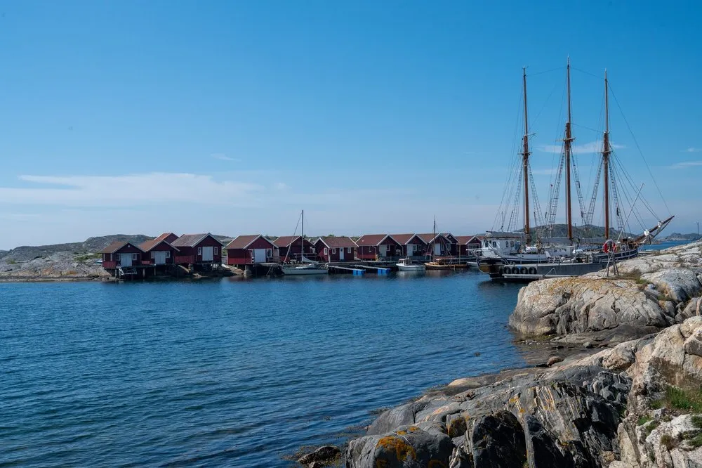Red boathouses on the shore of a bay, next to them a ship with three tall masts