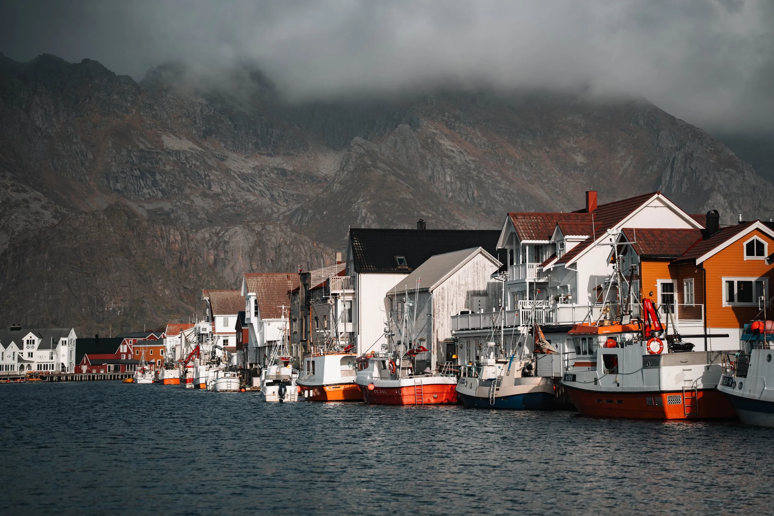 Fishing boats in the harbour of Henningsvær in front of the white and orange houses, dark clouds on the mountains above