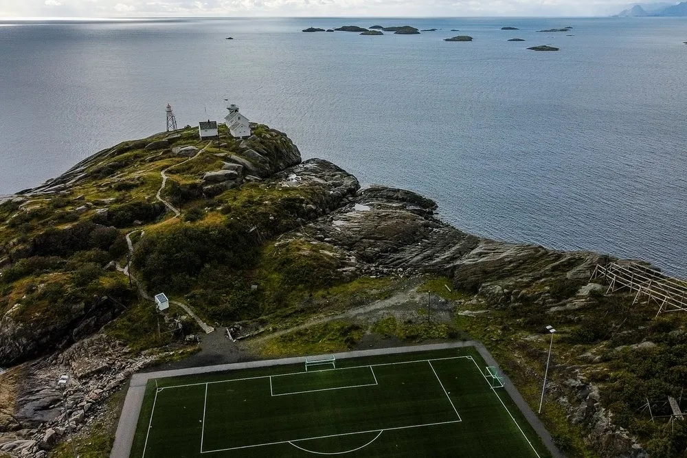 Der Fussballplatz von Henningsvær auf einer felsigen Landzunge am Meer, aus der Luft fotografiert, dahinter kleine Häuser und ein Mast
