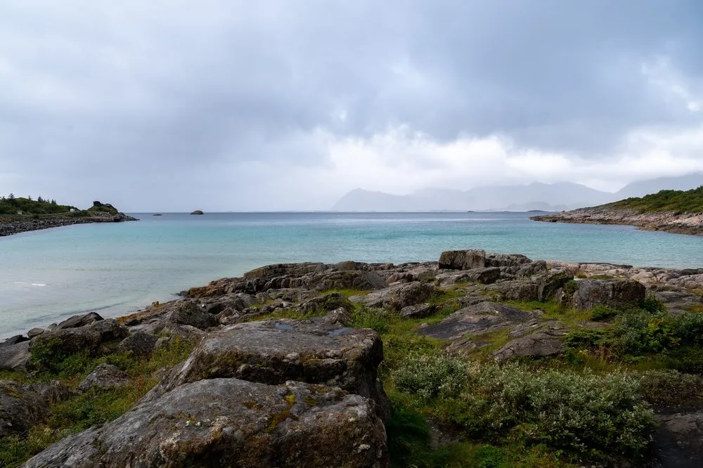 Rocky shore and turquoise water at Rørvikstranda under dark clouds, mountains in the haze in the background