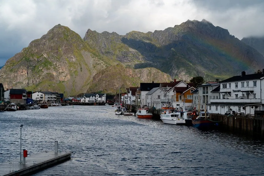 The harbour of Henningsvær with fishing boats and white houses, a mountain with a rainbow behind it