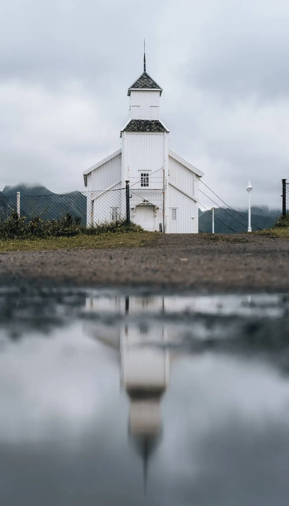 The white wooden church of Gimsøy with a pointed tower, its reflection lies in a puddle in the foreground