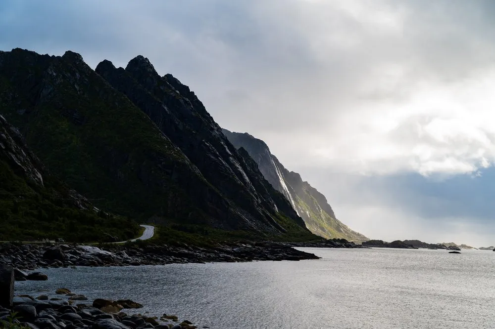 Steep dark mountainside by the water, a narrow road runs along its foot, sunlight falls through the clouds on the right