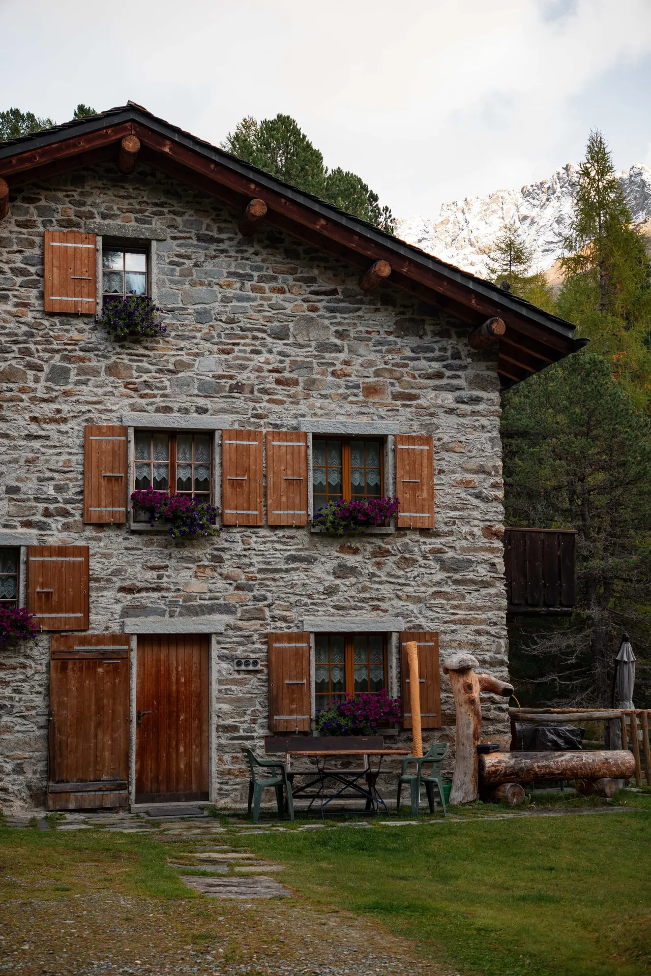 Ein Steinhaus in Lungacqua mit braunen Holzläden und violetten Blumen in den Fensterkästen, davor ein Gartentisch und ein Brunnen aus einem Baumstamm