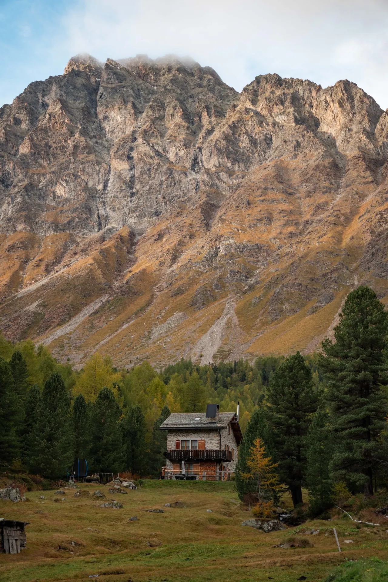 Ein Steinhaus mit Holzbalkon auf der Wiese von Lungacqua, dahinter eine hohe Felswand im Abendlicht