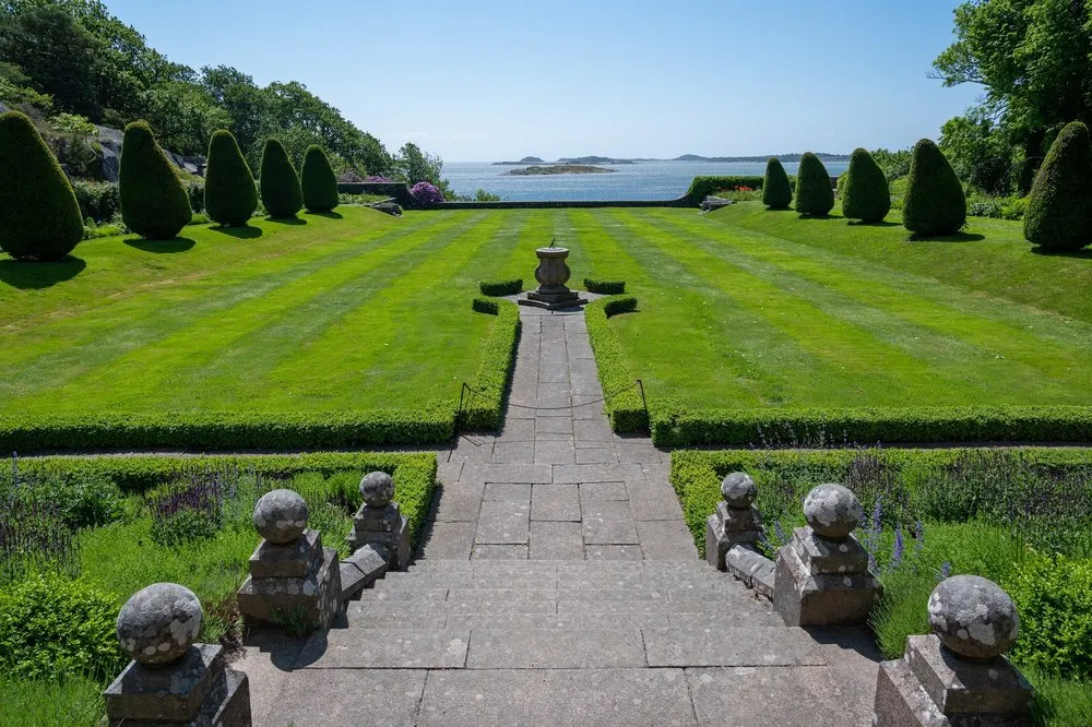 Der Schlossgarten von Tjolöholm mit Steintreppe, gemähtem Rasen und kegelförmig geschnittenen Bäumen, dahinter das Meer