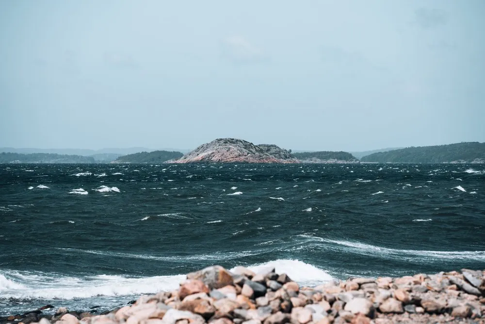 Dunkles, aufgewühltes Meer mit Schaumkronen vor einer felsigen Insel, im Vordergrund Steine