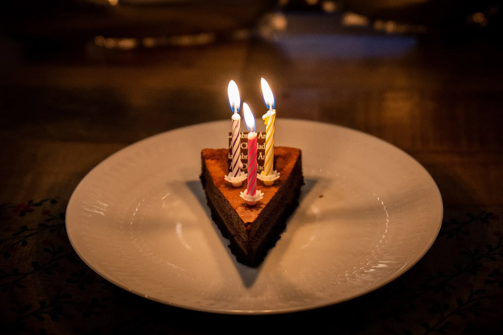 A slice of chocolate cake with three burning candles on a white plate