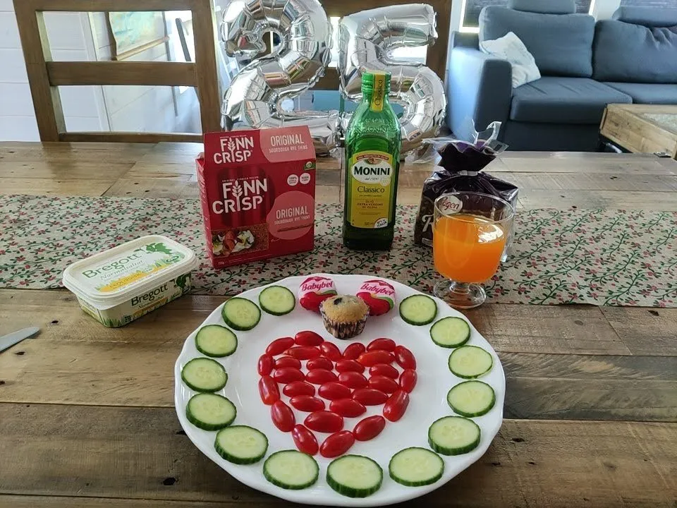 Breakfast table with a tomato heart and cucumber slices on the plate, a muffin, a packet of crispbread, butter, orange juice and silver balloons in the shape of a 25