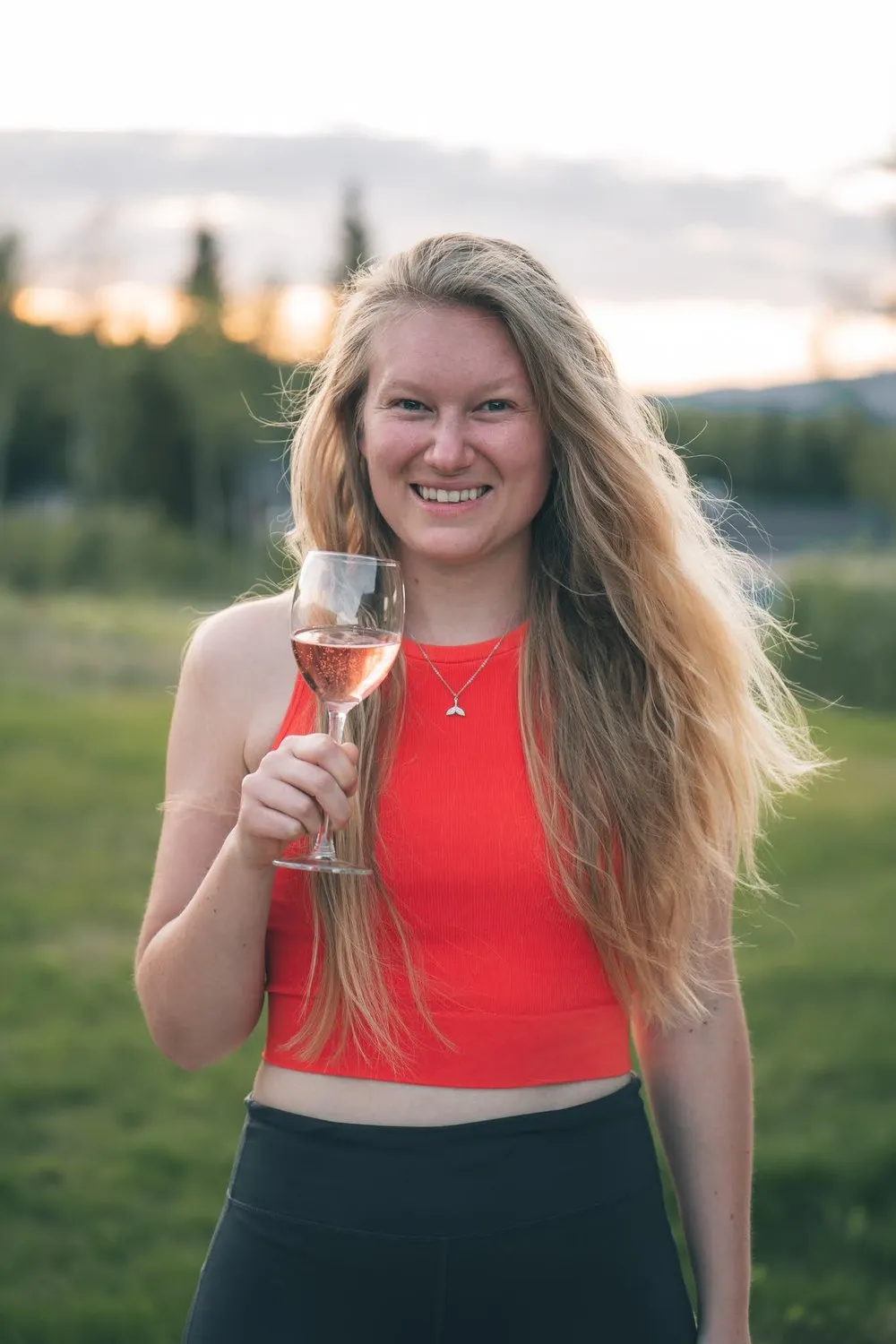 Annina in a red top laughs and holds a glass of rosé, behind her a meadow and forest in the evening light