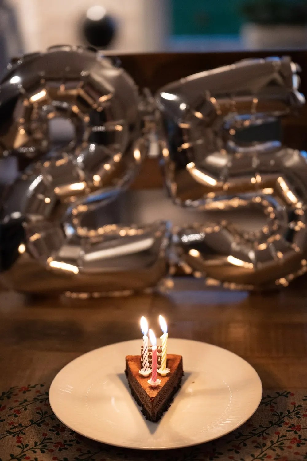 A slice of chocolate cake with three burning candles in front of silver balloons in the shape of a 25