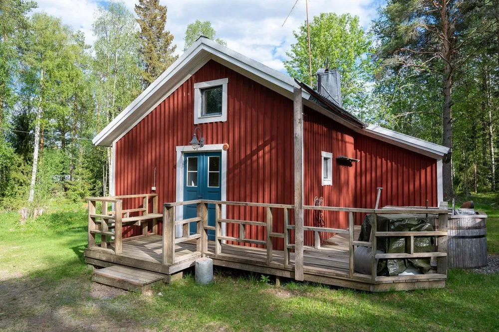 Red wooden house with a blue door and a wooden veranda, surrounded by birches and pines