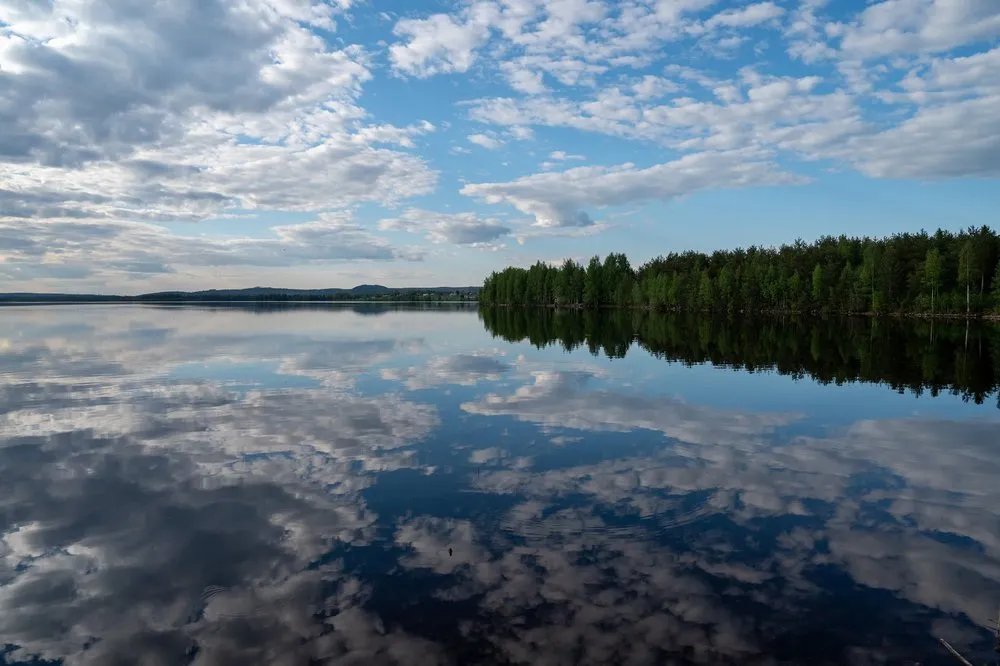 Calm lake with a wooded shore on the right, reflected in the water together with the clouds