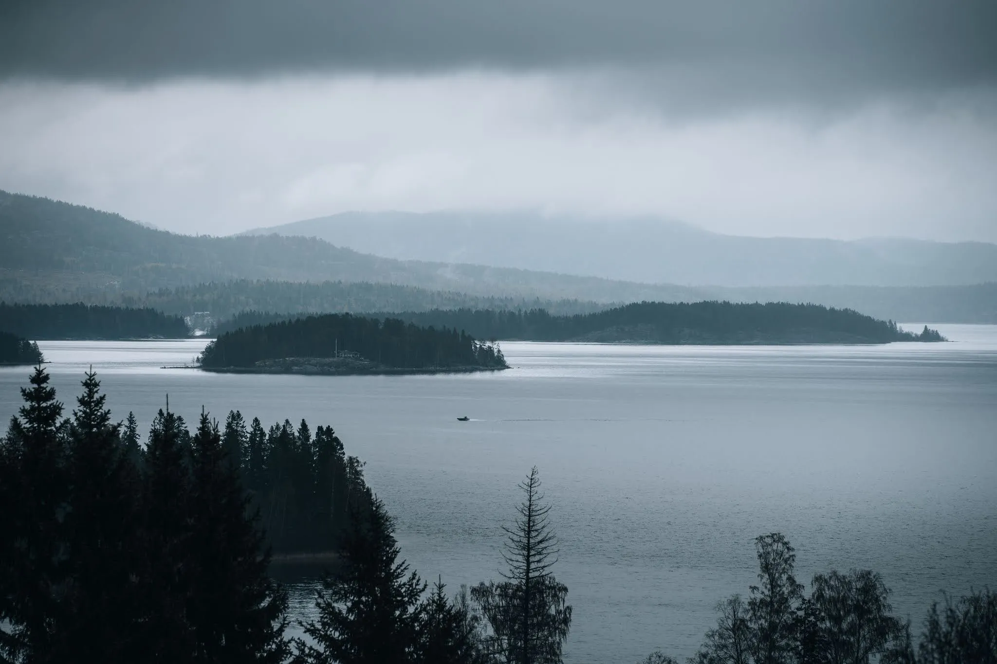 Wooded island in a grey expanse of water under low rain clouds, hazy hills behind