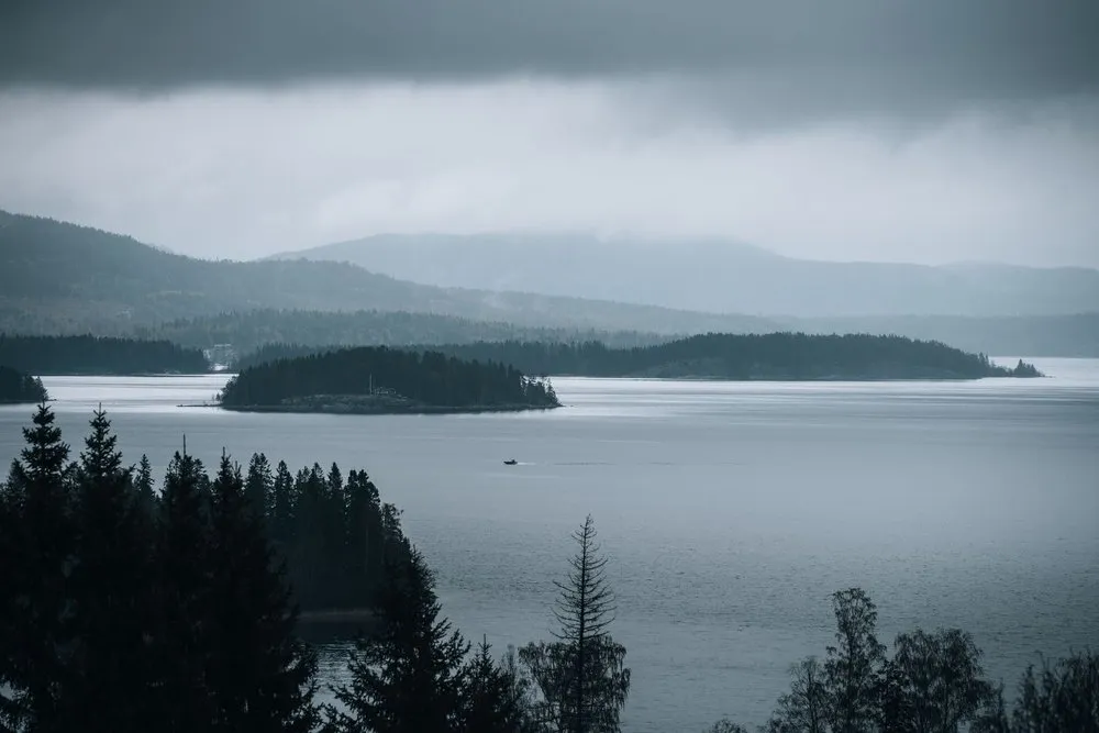 Bewaldete Insel in einer grauen Wasserfläche unter tiefen Regenwolken, dahinter Hügel im Dunst