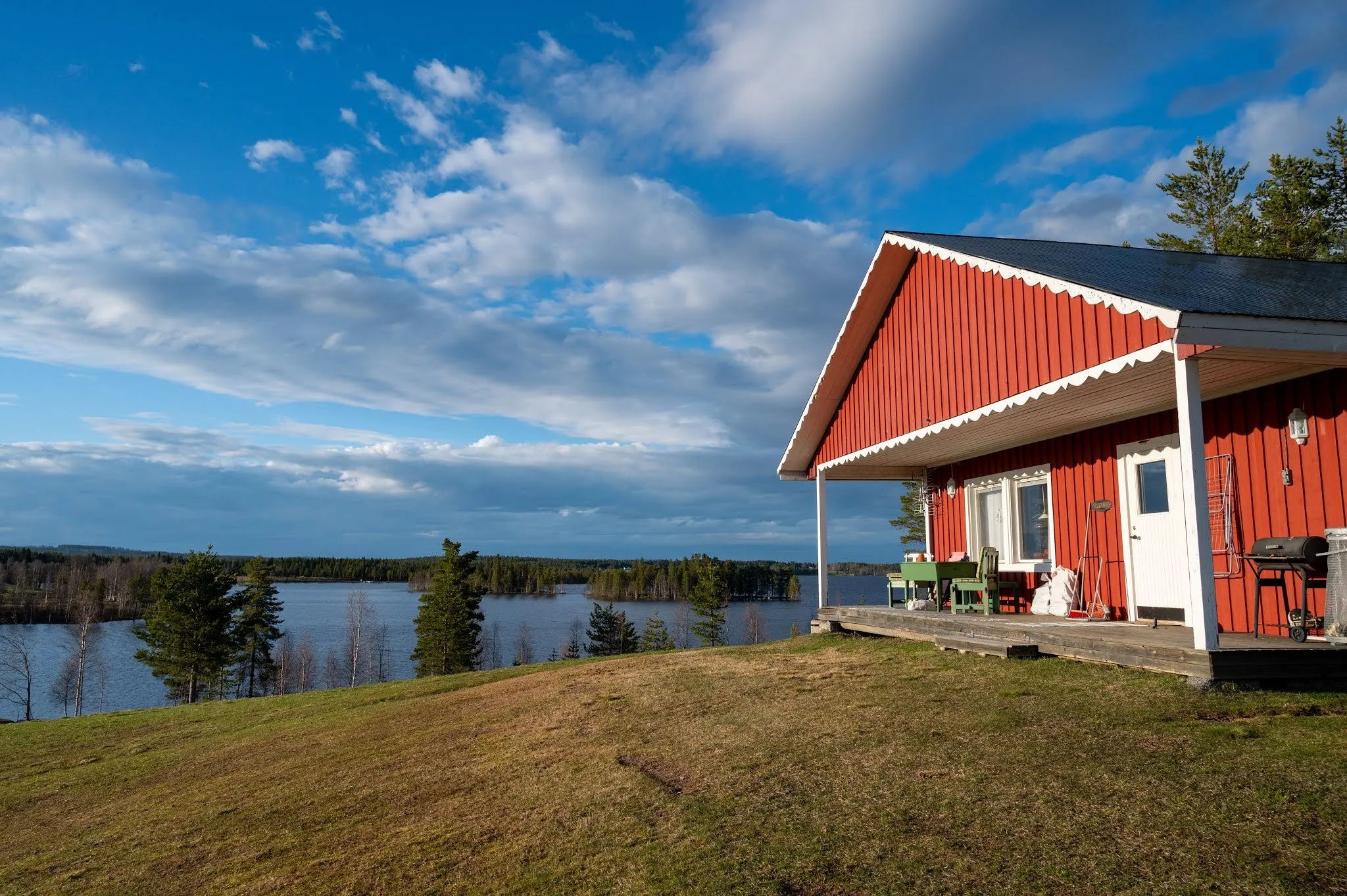 A red cottage with white trim stands on a meadow above a lake under a blue sky with clouds