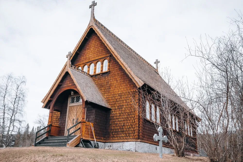 Die Holzkirche in Kvikkmokk mit Schindelfassade und einem Steinkreuz davor