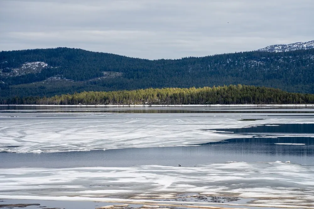 Ein teils zugefrorener See mit Eisschollen vor bewaldeten Hügeln mit einzelnen Schneeflecken