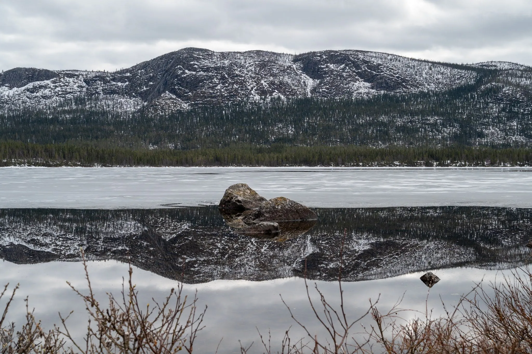 A boulder juts out of the mirror-smooth water, with the reflection of a snow-covered mountain slope in it