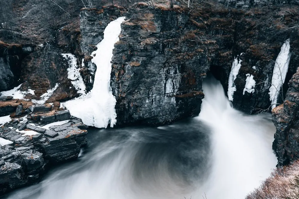 Der Wasserfall im Abisko Nationalpark stürzt zwischen dunklen Felswänden mit Eisresten in ein weiss schäumendes Becken