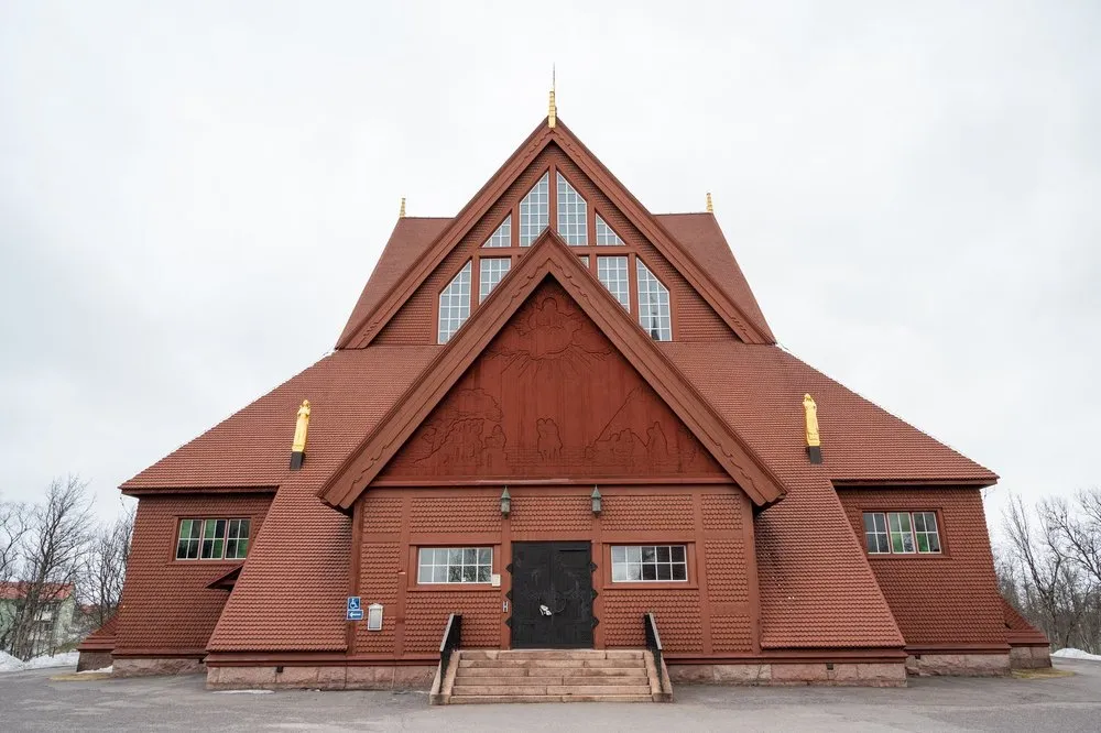 Die rotbraune Holzkirche von Kiruna mit steilen Giebeln, Schindeldach und goldenen Figuren auf den Firsten unter grauem Himmel