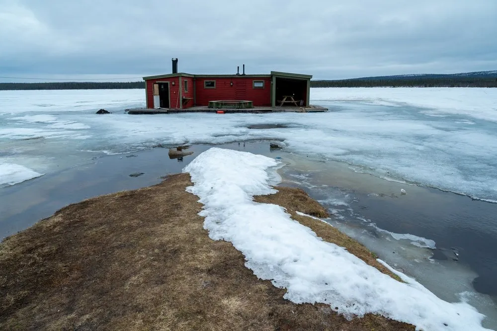 Die rote Sauna auf dem Eis des Sees beim Campingplatz, davor ein Streifen braunes Gras mit Schneeresten und Schmelzwasser