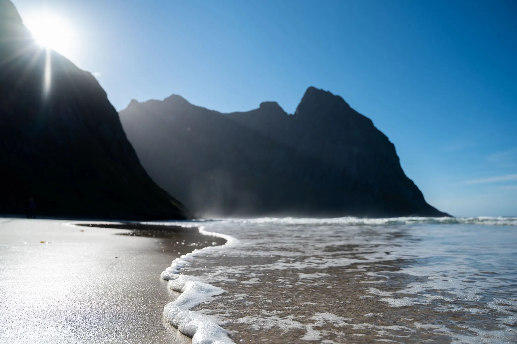 Backlight over a dark rock wall, in the foreground a wave with foam runs over wet sand