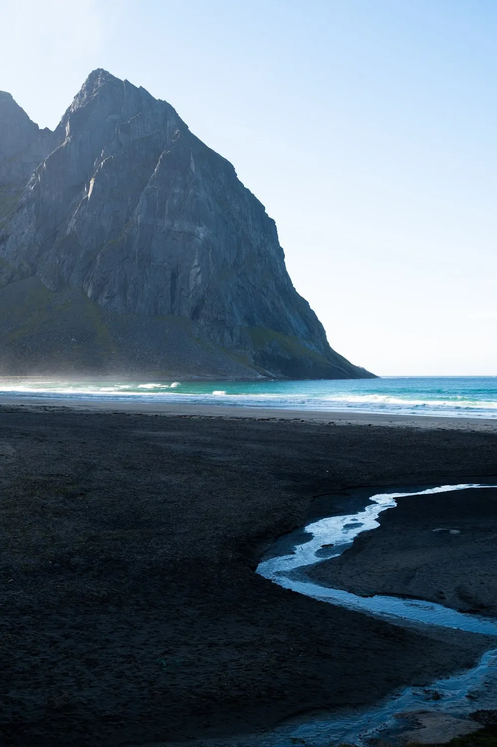A stream winds across the dark sand, a rock wall towers behind it