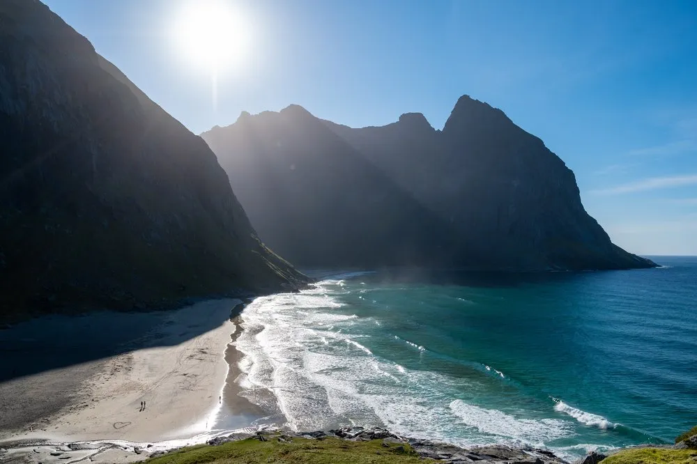 Sandy beach with surf and a few people beneath the rock walls, the sun above