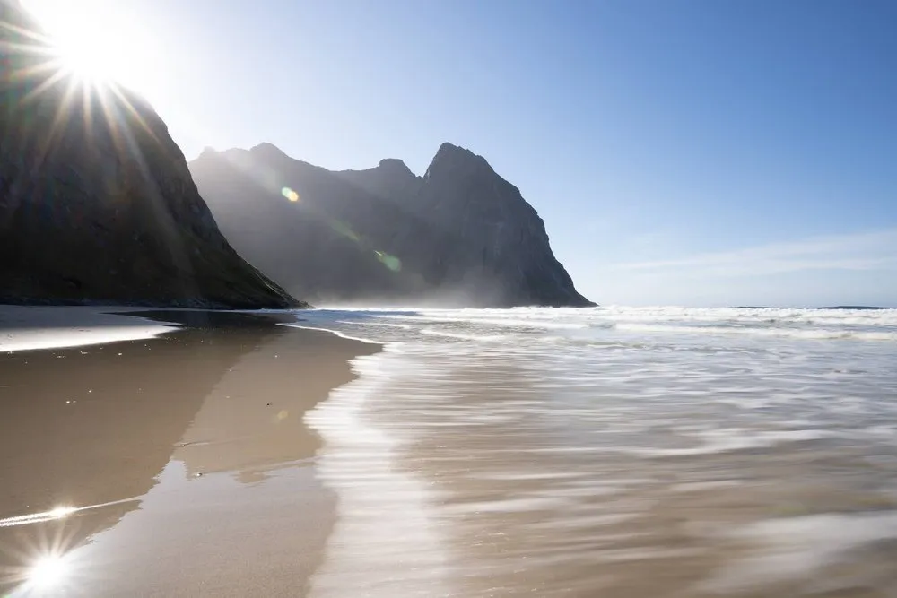 Waves running out in a long exposure on wet sand, the sun shining from behind the rock