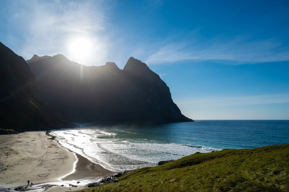 Looking across a green slope edge down to the beach, the sun directly above the mountain ridge
