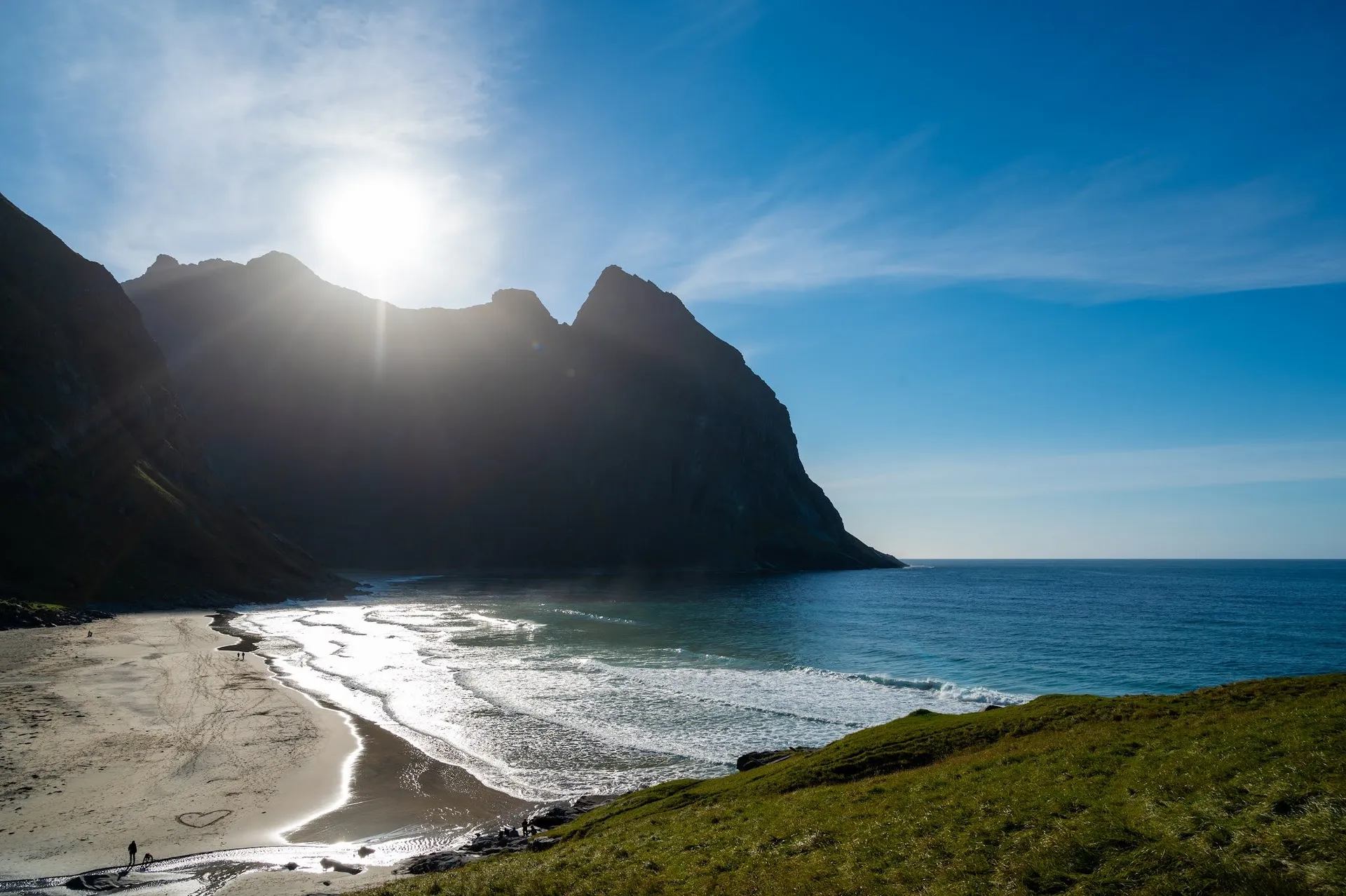 Blick über eine grüne Hangkante auf den Strand, die Sonne steht direkt über dem Bergkamm