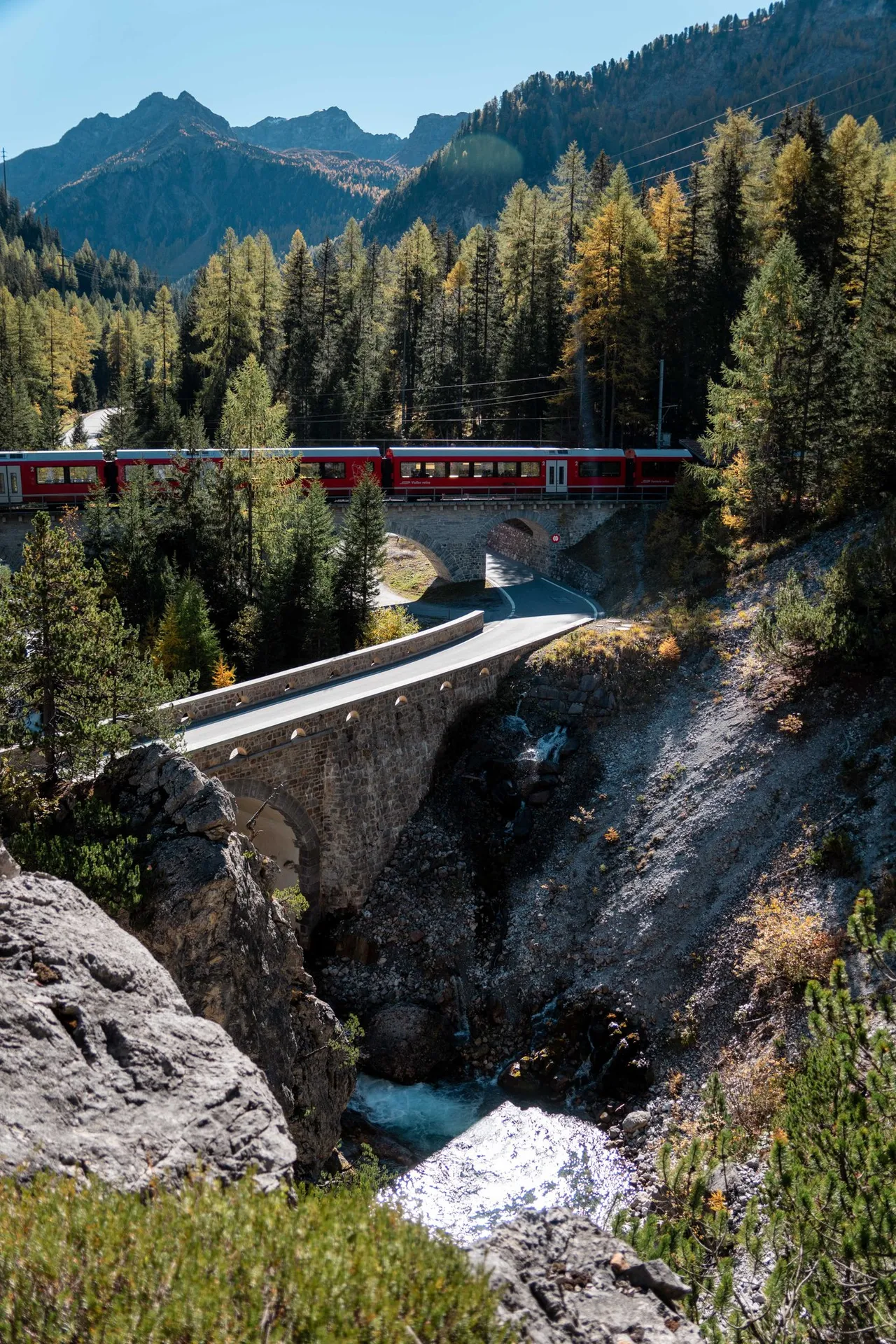 Ein roter Zug auf einem niedrigen Steinviadukt, unter dem die Strasse hindurchführt, davor eine Strassenbrücke über die Albula