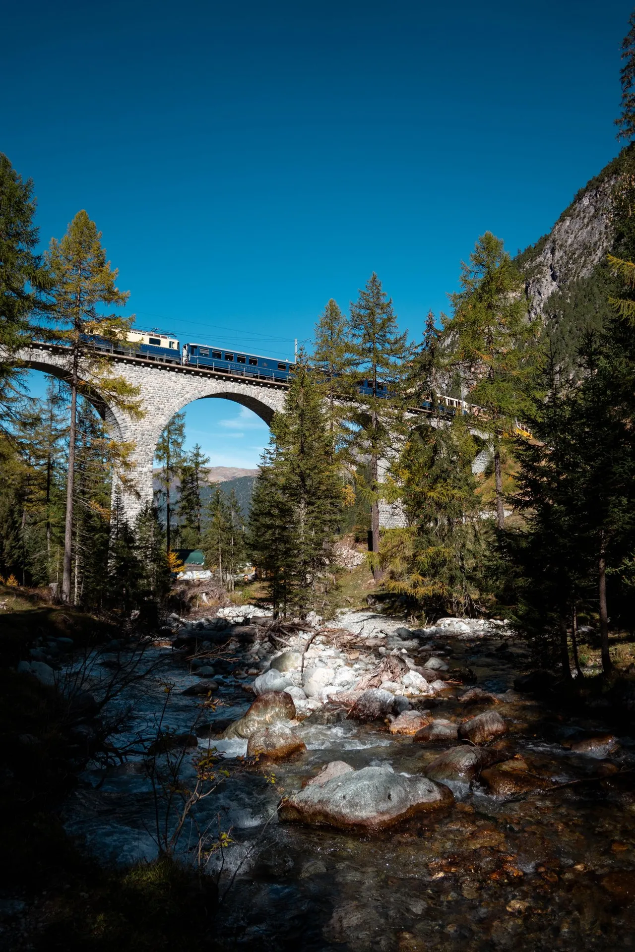 Die blau-elfenbeinfarbene Lok, der Speisewagen Gourmino und weitere blaue Wagen auf dem Viadukt über dem Bach