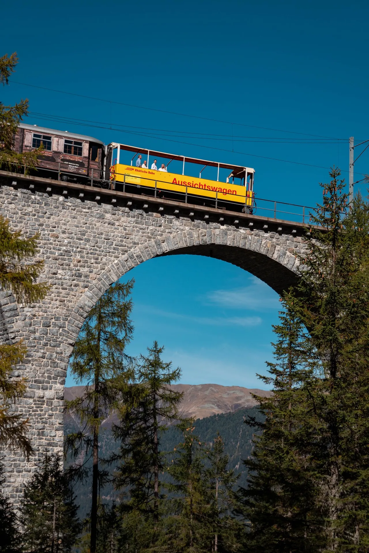Ein Holzwagen und der gelbe Aussichtswagen der Rhätischen Bahn über dem Bogen des Viadukts