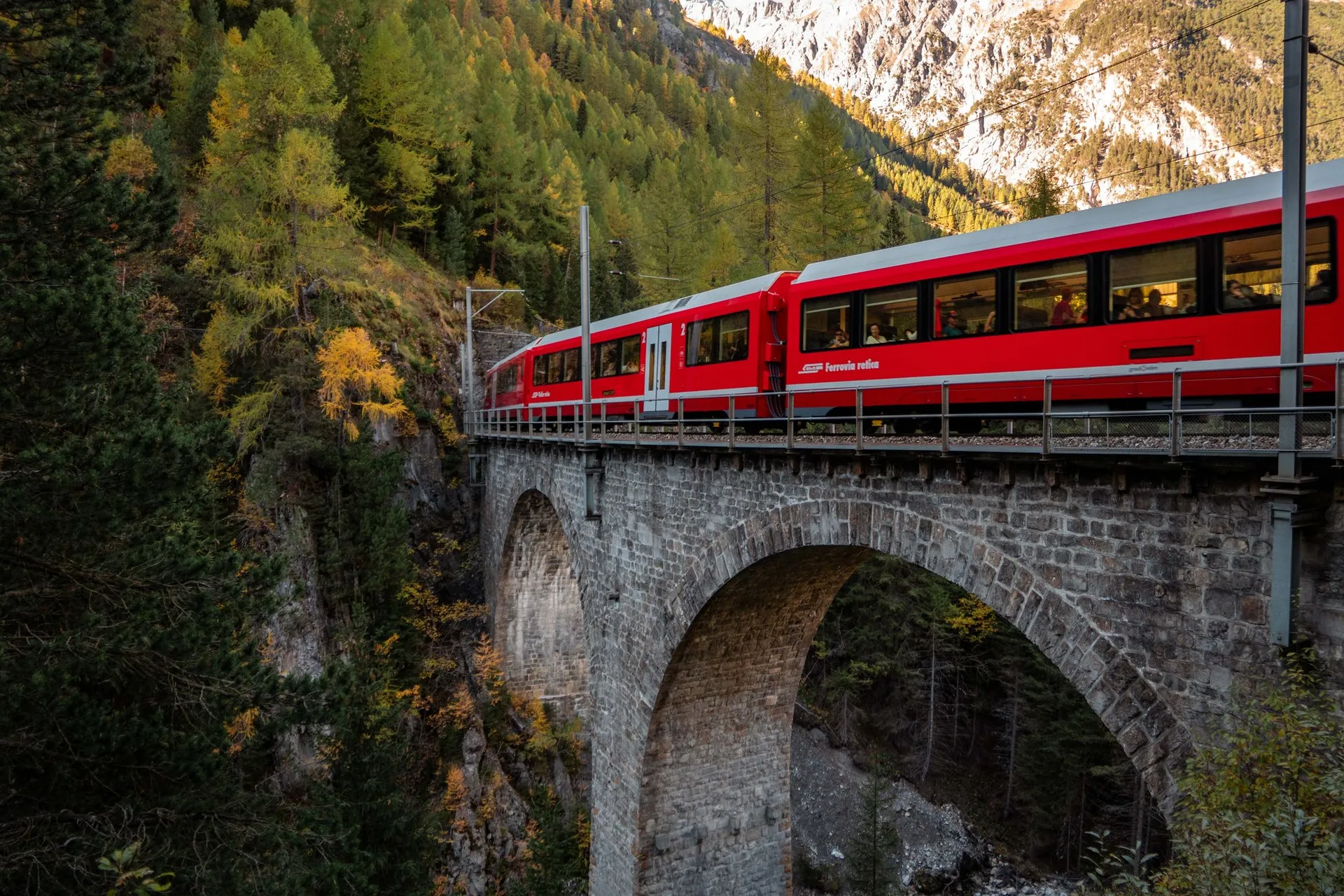 Ein roter Zug der Rhätischen Bahn auf einem steinernen Bogenviadukt über einer Schlucht, dahinter gelbe Lärchen