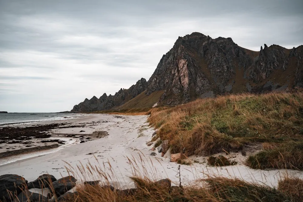 Ein weisser Sandstrand bei Bleik mit trockenem Gras, dahinter ein gezackter dunkler Felsgrat unter grauen Wolken