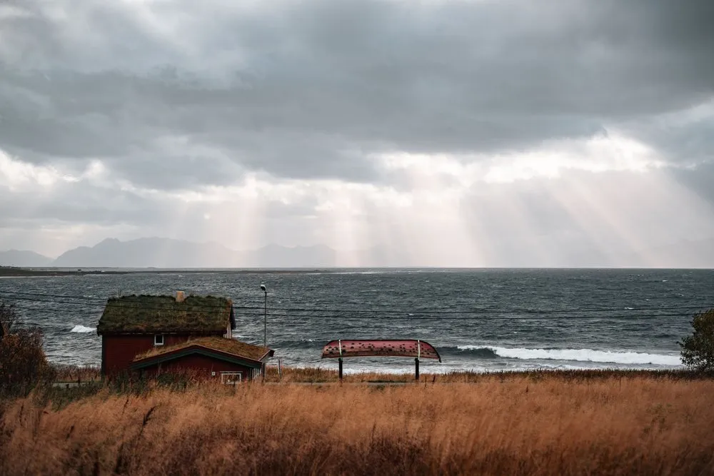 Sunbeams breaking through dark storm clouds over the rough sea near Nøss, a house with a grass roof and dry grass in front