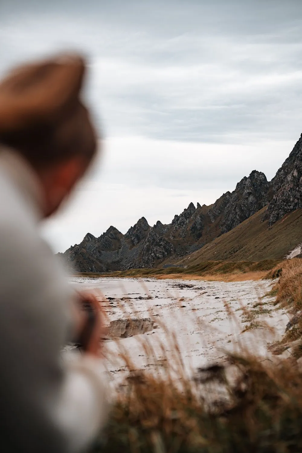 Annina in a hat, out of focus in the foreground on the beach at Bleik, the jagged ridge and the pale sand behind