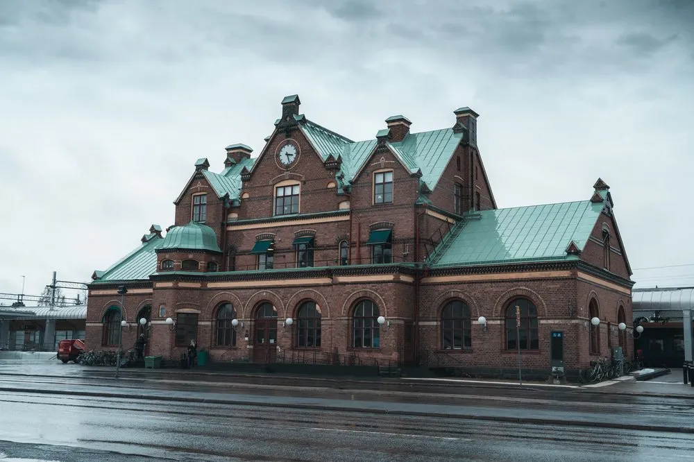 Ein Backsteinbau mit grünem Kupferdach, Türmchen und Uhr an einer regennassen Strasse unter grauem Himmel