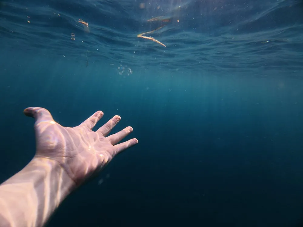 Eine Hand streckt sich unter Wasser ins tiefe Blau, darüber das Licht der Oberfläche