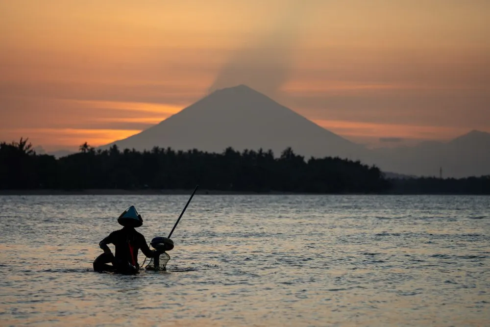 A fisherman in a hat wades through shallow water, a volcanic cone stands out against an orange sky behind him